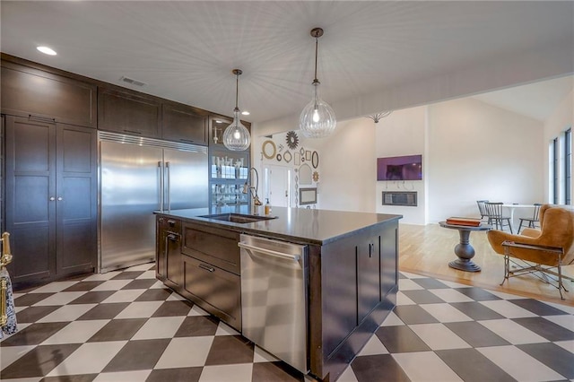 kitchen with dark floors, a center island with sink, stainless steel appliances, hanging light fixtures, and a sink