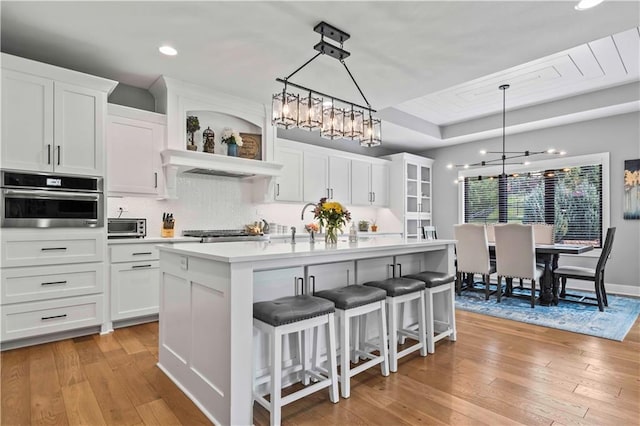 kitchen with appliances with stainless steel finishes, light wood-type flooring, an island with sink, white cabinetry, and decorative light fixtures