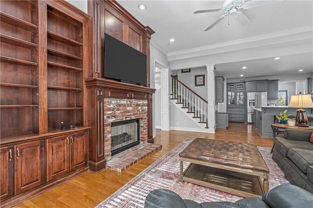 living room featuring a ceiling fan, ornamental molding, stairs, light wood-style floors, and a fireplace