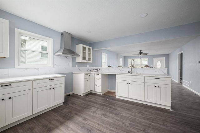 kitchen with dark wood-type flooring, kitchen peninsula, wall chimney exhaust hood, sink, and white cabinetry