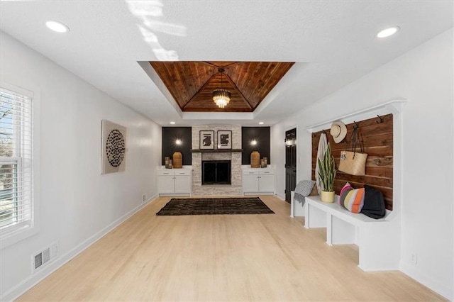 living area featuring baseboards, visible vents, light wood finished floors, a tray ceiling, and a stone fireplace