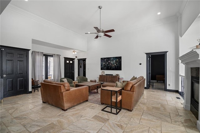 living area featuring baseboards, a ceiling fan, a glass covered fireplace, ornamental molding, and a high ceiling