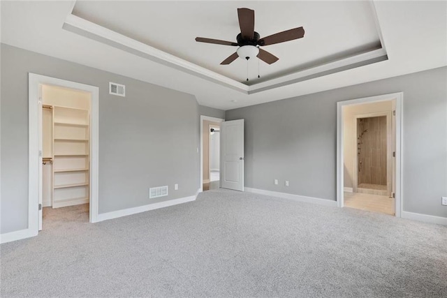 unfurnished bedroom featuring light carpet, ensuite bathroom, ceiling fan, a spacious closet, and a tray ceiling