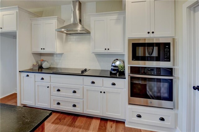 kitchen featuring wall chimney range hood, built in microwave, white cabinets, black electric cooktop, and stainless steel oven