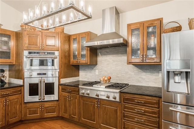 kitchen with appliances with stainless steel finishes, light hardwood / wood-style flooring, dark stone counters, and wall chimney range hood