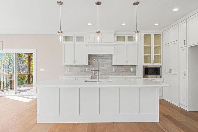 kitchen with light wood finished floors, a center island with sink, light countertops, decorative backsplash, and a sink