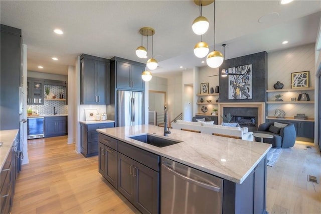 kitchen with a large fireplace, stainless steel appliances, a kitchen island with sink, sink, and light hardwood / wood-style floors