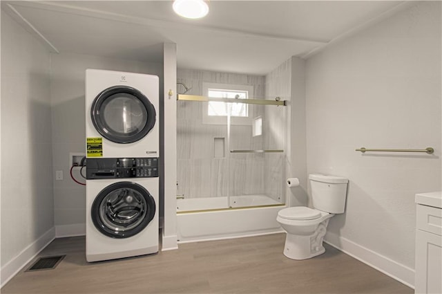 full bathroom with stacked washing maching and dryer, bath / shower combo with glass door, vanity, wood-type flooring, and toilet