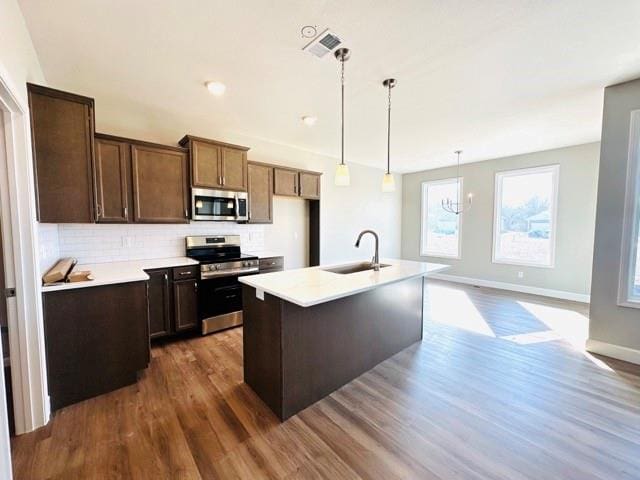 kitchen featuring sink, backsplash, stainless steel appliances, an island with sink, and decorative light fixtures