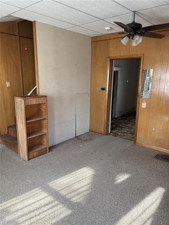 empty room featuring a drop ceiling, wooden walls, ceiling fan, and carpet flooring