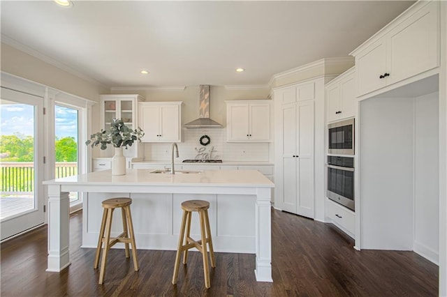 kitchen featuring sink, wall chimney range hood, oven, an island with sink, and white cabinets