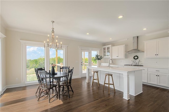 kitchen featuring an inviting chandelier, a sink, wall chimney exhaust hood, a center island with sink, and dark wood-style flooring