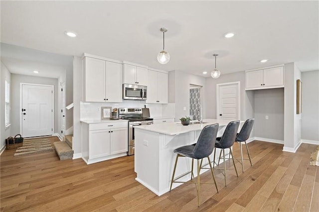 kitchen with a kitchen island with sink, white cabinets, light wood-type flooring, decorative light fixtures, and stainless steel appliances