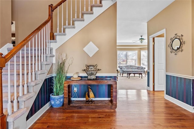 foyer entrance featuring a wainscoted wall, stairway, ceiling fan, wood finished floors, and baseboards