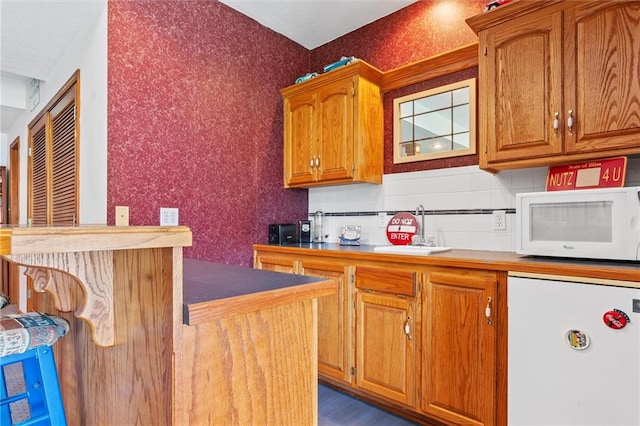 kitchen with wallpapered walls, brown cabinetry, white microwave, fridge, and a sink