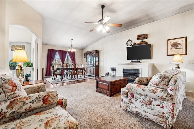 carpeted living room featuring a fireplace, vaulted ceiling, ceiling fan with notable chandelier, and arched walkways