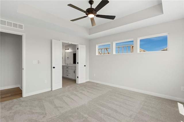 unfurnished bedroom featuring ceiling fan, a tray ceiling, ensuite bathroom, and light carpet