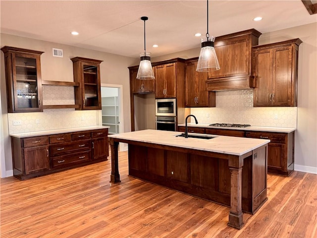 kitchen with sink, hanging light fixtures, stainless steel appliances, an island with sink, and light wood-type flooring