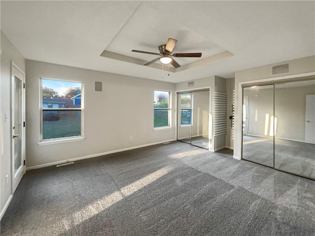 unfurnished bedroom with a tray ceiling, ceiling fan, dark carpet, and two closets