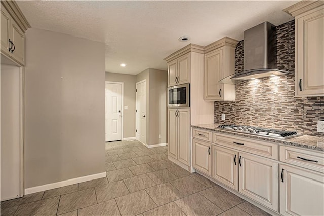 kitchen with cream cabinetry, light stone counters, wall chimney range hood, and appliances with stainless steel finishes