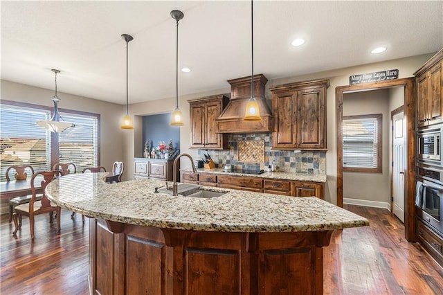 kitchen with sink, appliances with stainless steel finishes, premium range hood, hanging light fixtures, and an island with sink