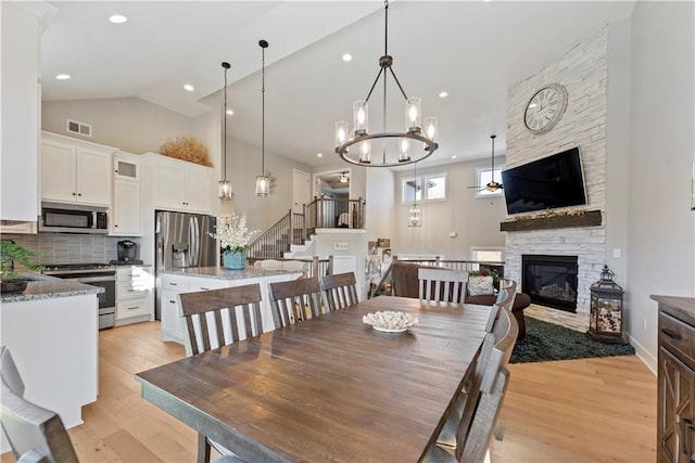 dining space featuring a stone fireplace, high vaulted ceiling, light hardwood / wood-style floors, and ceiling fan with notable chandelier