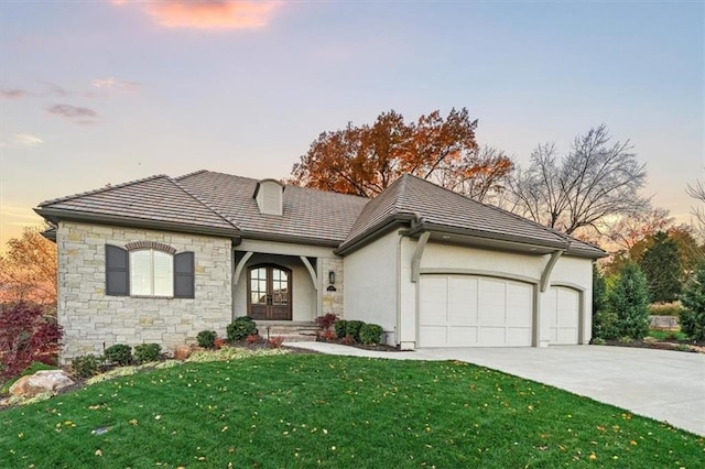 french country inspired facade featuring a garage, driveway, a yard, stone siding, and french doors