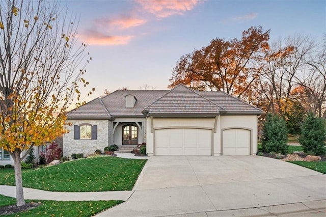 french country inspired facade with a garage, stone siding, concrete driveway, stucco siding, and a front yard