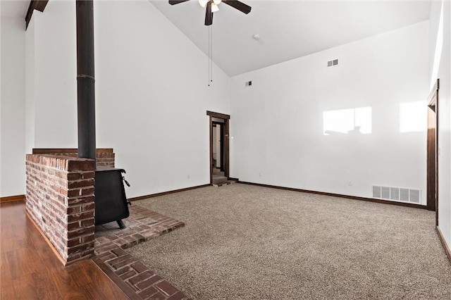 unfurnished living room with dark colored carpet, high vaulted ceiling, a wood stove, and ceiling fan