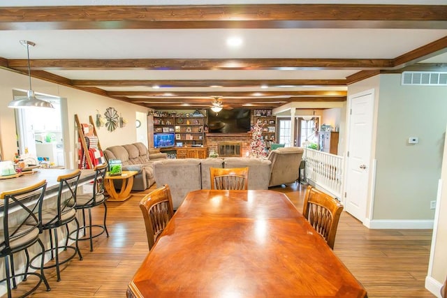 dining area featuring beamed ceiling, light hardwood / wood-style floors, a brick fireplace, and ceiling fan