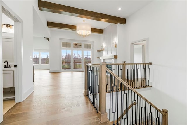 corridor featuring light wood finished floors, a sink, an upstairs landing, beamed ceiling, and baseboards