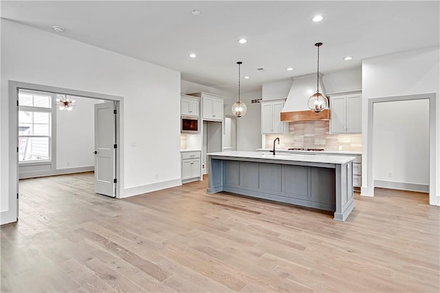 kitchen with light hardwood / wood-style flooring, pendant lighting, decorative backsplash, a center island with sink, and custom exhaust hood