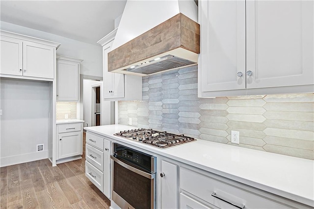 kitchen featuring backsplash, light hardwood / wood-style flooring, custom range hood, appliances with stainless steel finishes, and white cabinetry