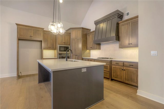 kitchen with sink, a kitchen island with sink, custom range hood, stainless steel appliances, and light hardwood / wood-style floors