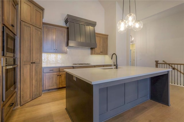 kitchen with sink, hanging light fixtures, light wood-type flooring, appliances with stainless steel finishes, and an island with sink