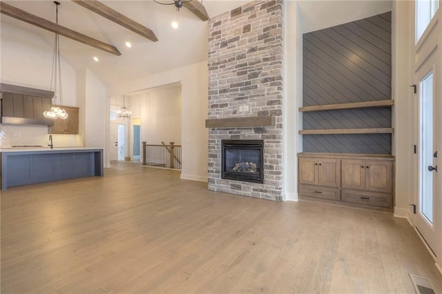 unfurnished living room with a stone fireplace, high vaulted ceiling, beamed ceiling, light hardwood / wood-style floors, and an inviting chandelier