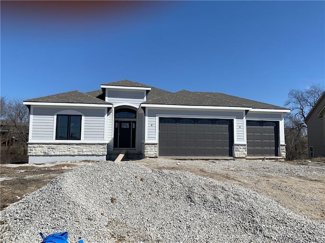 prairie-style home with gravel driveway, a garage, stone siding, and roof with shingles