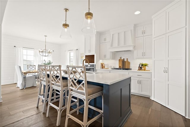 kitchen with custom exhaust hood, an island with sink, cooktop, light countertops, and white cabinets