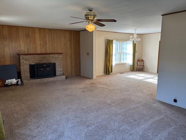 unfurnished living room featuring ceiling fan with notable chandelier, wood walls, light colored carpet, and a brick fireplace