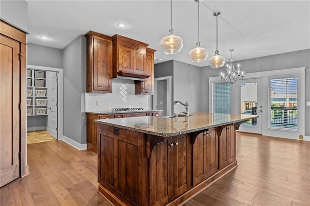 kitchen with sink, a center island with sink, stone countertops, light hardwood / wood-style floors, and hanging light fixtures