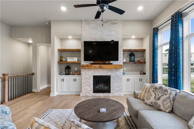 living room featuring built in shelves, light wood-type flooring, a stone fireplace, and ceiling fan