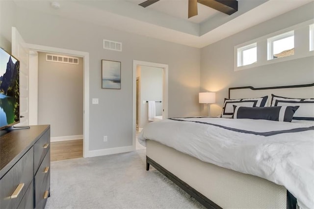 bedroom with ceiling fan, light colored carpet, ensuite bathroom, and a raised ceiling