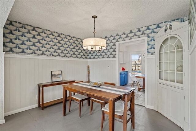 dining room featuring a textured ceiling, a wainscoted wall, baseboards, finished concrete flooring, and wallpapered walls