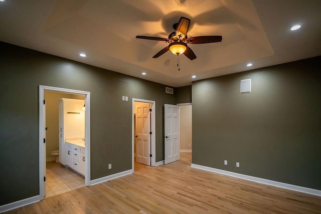 unfurnished bedroom with ensuite bathroom, a raised ceiling, ceiling fan, and light wood-type flooring