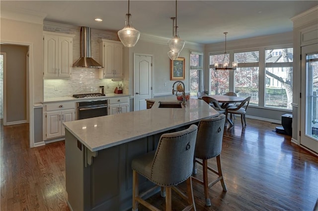 kitchen with wall oven, wall chimney exhaust hood, sink, a large island with sink, and white cabinets