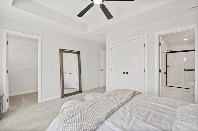 carpeted bedroom featuring a tray ceiling, ensuite bath, ceiling fan, and multiple closets