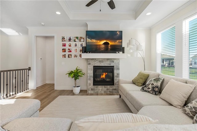 living room featuring a raised ceiling, ceiling fan, crown molding, light hardwood / wood-style flooring, and a stone fireplace