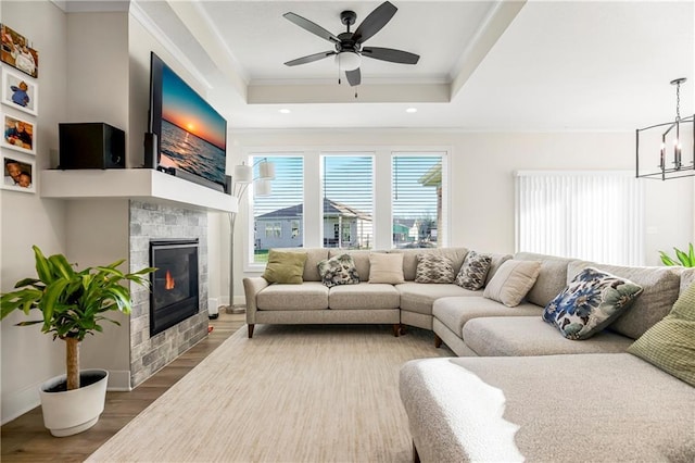 living room featuring ceiling fan with notable chandelier, a raised ceiling, crown molding, wood-type flooring, and a tiled fireplace