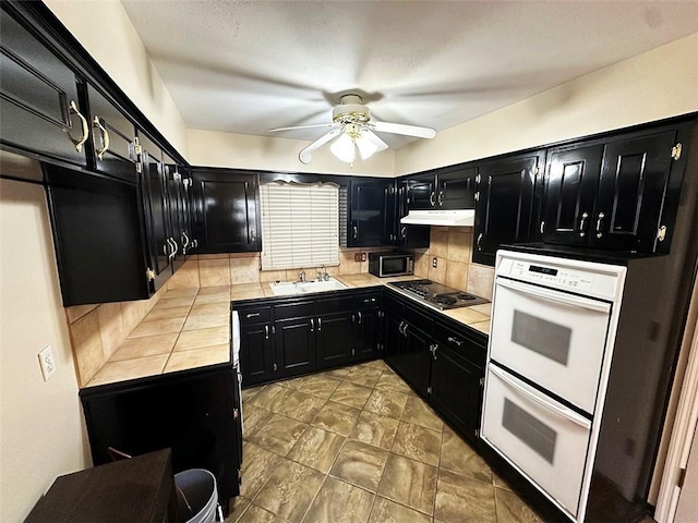 kitchen with white double oven, backsplash, sink, ceiling fan, and stainless steel gas cooktop