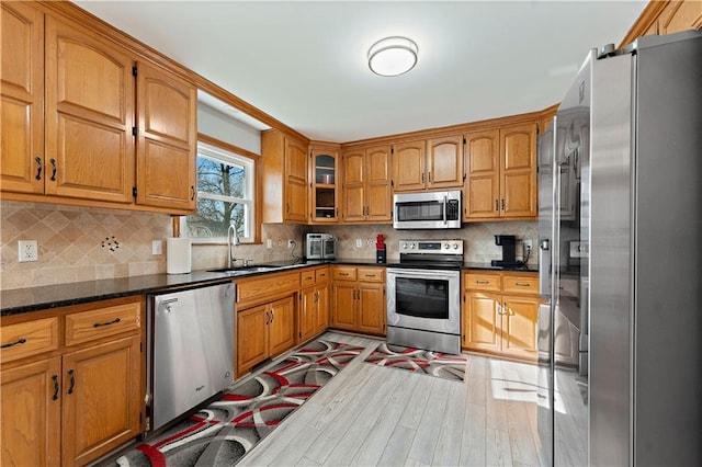 kitchen with sink, light wood-type flooring, dark stone countertops, appliances with stainless steel finishes, and backsplash